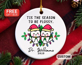 May include: White ceramic Christmas ornament with the text "TIS THE SEASON TO BE FLOSSY." Features two cartoon teeth wearing Santa hats, surrounded by Christmas lights and holly. Includes the name "Dr. Williams" and the year "2025".