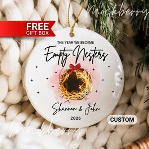 May include: A white ceramic Christmas ornament with a gold string for hanging. The ornament features the text "The Year We Became Empty Nesters" and the names "Shannon & John" with the year 2026. A nest illustration is in the center.