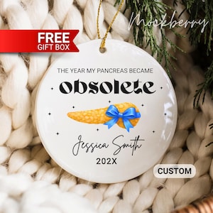May include: White ceramic ornament with gold string, featuring the text "THE YEAR MY PANCREAS BECAME OBSOLETE" and a pancreas illustration with a blue bow. Includes a name and year.