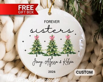 May include: White ceramic Christmas ornament with gold string, featuring the text "FOREVER sisters" above three green Christmas trees with pink bows. The names "Jenny, Alyssa & Kelsea" and the year "2026" are also printed on the ornament.
