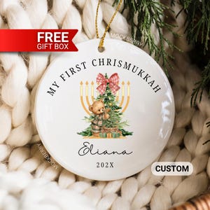 May include: A round white ceramic ornament featuring the text MY FIRST CHRISMUKKAH, the name Eliana, and the year 202X. The design includes a brown teddy bear decorating a green Christmas tree, a yellow menorah, and a red and white plaid bow.