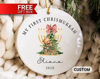 May include: A round white ceramic ornament featuring the text MY FIRST CHRISMUKKAH, the name Eliana, and the year 202X. The design includes a brown teddy bear decorating a green Christmas tree, a yellow menorah, and a red and white plaid bow.