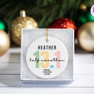 May include: A white ceramic ornament with the text "HEATHER 13.1 half marathon CHICAGO 11/08/2025 2:25:05" in various colors. The ornament is in a white box, with gold and red Christmas ornaments in the background.