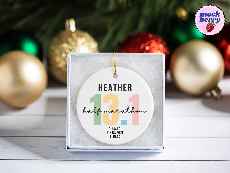 May include: A white ceramic ornament with the text "HEATHER 13.1 half marathon CHICAGO 11/08/2025 2:25:05" in various colors. The ornament is in a white box, with gold and red Christmas ornaments in the background.