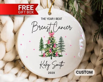 May include: A round white ceramic ornament featuring the text THE YEAR I BEAT Breast Cancer, a central Christmas tree decorated with pink ribbons, the name Kaley Smith, and the year 202X, suspended by a gold string against a white knit background.
