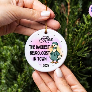Puede incluir: Un adorno de cer&aacute;mica blanca con una cuerda dorada, sostenido por manos. El adorno presenta el texto "Alex" encima de "THE BADDEST NEUROLOGIST IN TOWN" y "2025". Un pato de dibujos animados con uniforme tambi&eacute;n est&aacute; en el adorno.