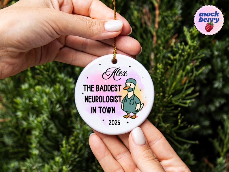 Puede incluir: Un adorno de cer&aacute;mica blanca con una cuerda dorada, sostenido por manos. El adorno presenta el texto "Alex" encima de "THE BADDEST NEUROLOGIST IN TOWN" y "2025". Un pato de dibujos animados con uniforme tambi&eacute;n est&aacute; en el adorno.