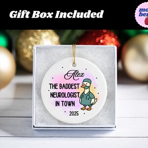 Puede incluir: Adorno de cer&aacute;mica blanca en una caja de regalo. El adorno presenta un pato de dibujos animados con uniforme m&eacute;dico, con el texto "Alex, The Baddest Neurologist in Town, 2025". El fondo tiene un efecto acuarela con tonos rosas y amarillos. Se adjunta un cord&oacute;n dorado para colgar.