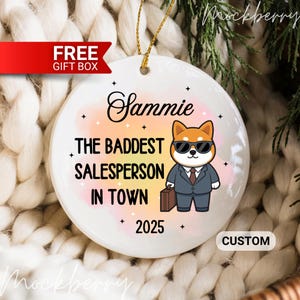 May include: A white ceramic ornament with a gold string for hanging. The ornament features the name "Sammie" and the text "THE BADDEST SALESPERSON IN TOWN 2025". A cartoon Shiba Inu in a suit and sunglasses is also on the ornament.