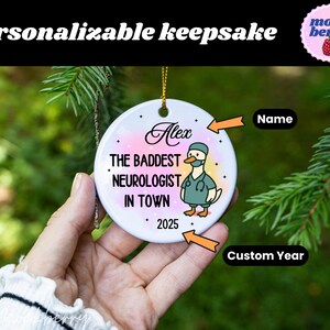 Puede incluir: Un adorno de recuerdo personalizable con el texto "Personalizable keepsake". El adorno presenta el nombre "Alex" y la frase "THE BADDEST NEUROLOGIST IN TOWN" con un pato de dibujos animados con uniforme. Tambi&eacute;n se muestra el a&ntilde;o "2025".