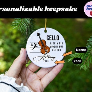 May include: A white ceramic ornament with a cello illustration and musical notes. Text includes "CELLO," "LIKE A BIG VIOLIN BUT BETTER," a name, and the year 2025. The ornament is hanging from a gold string.