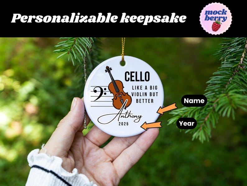 May include: A white ceramic ornament with a cello illustration and musical notes. Text includes "CELLO," "LIKE A BIG VIOLIN BUT BETTER," a name, and the year 2025. The ornament is hanging from a gold string.