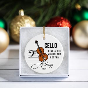May include: A white ceramic Christmas ornament with a cello illustration, musical notes, and the text "CELLO LIKE A BIG VIOLIN BUT BETTER". Includes a name and the year 2025. The ornament is in a white gift box.
