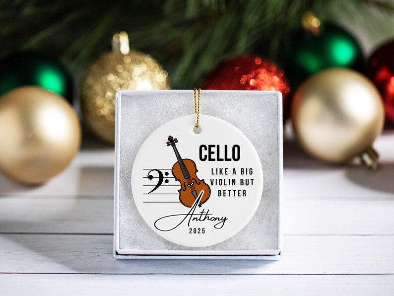 May include: A white ceramic Christmas ornament with a cello illustration, musical notes, and the text "CELLO LIKE A BIG VIOLIN BUT BETTER". Includes a name and the year 2025. The ornament is in a white gift box.