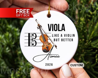 May include: A round white ceramic ornament featuring a brown viola illustration, a musical staff with a C-clef, and the text: VIOLA LIKE A VIOLIN BUT BETTER, Aimee, and 2026. The ornament hangs from a gold string and includes a hole at the top.