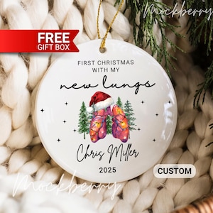 May include: White ceramic Christmas ornament with gold string. The ornament features the text "First Christmas with my new lungs" and a watercolor illustration of lungs wearing a Santa hat. The name "Chris Miller" and the year "2025" are also included.