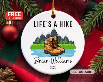 May include: White ceramic ornament with the text "LIFE'S A HIKE" and a graphic of hiking boots, mountains, and water. The name "Brian Williams" and the year "2025" are also printed on the ornament. A gold string is attached for hanging.