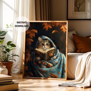 Könnte beinhalten: Ein gerahmter Druck, der eine Katze beim Lesen eines Buches zeigt, eingehüllt in eine blaue Decke mit orangefarbenen Blumenmustern. Die Katze hat braunes und beiges Fell und trägt eine blaue Kapuze. Der Druck ist mit "INSTANT DOWNLOAD" beschriftet.