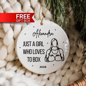 May include: White ceramic ornament with gold string for hanging. The ornament features the name "Alexandra", the phrase "JUST A GIRL WHO LOVES TO BOX", a boxing figure, and the year "2025". A red banner with "FREE GIFT BOX" is in the upper left.