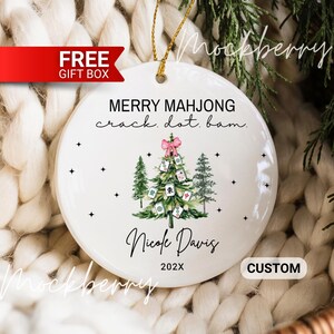 May include: A round white ceramic ornament featuring a Christmas tree decorated with mahjong tiles, a pink bow, and green pine trees. The text reads MERRY MAHJONG crack. dot. bam. Nicole Davis 202X. It includes a gold hanging string.