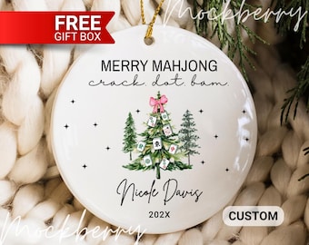 May include: A round white ceramic ornament featuring a Christmas tree decorated with mahjong tiles, a pink bow, and green pine trees. The text reads MERRY MAHJONG crack. dot. bam. Nicole Davis 202X. It includes a gold hanging string.