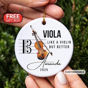 May include: White ceramic ornament with a violin illustration and the text "VIOLA LIKE A VIOLIN BUT BETTER". Includes a musical staff, a bow, and the name "Amanda" with the year "2025".