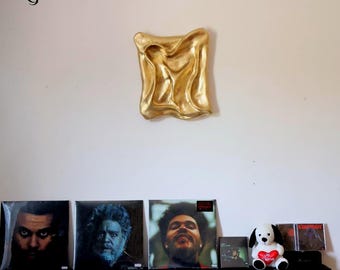 May include: A gold-coloured, abstract wall sculpture with a textured surface is mounted on a white wall. Below, a black shelf displays several vinyl record albums and a stuffed dog holding a heart.