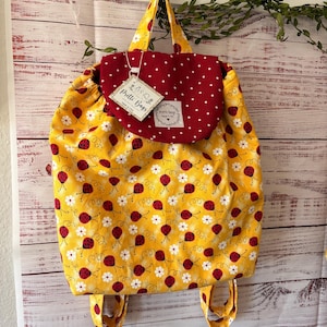 Peut inclure: Un sac &agrave; dos &agrave; cordon en tissu jaune avec un motif de coccinelles rouges et de marguerites blanches. Le sac poss&egrave;de un rabat rouge fonc&eacute; &agrave; pois blancs et une &eacute;tiquette indiquant Pretti Bags Made in USA. Il comprend deux anses et des bretelles.