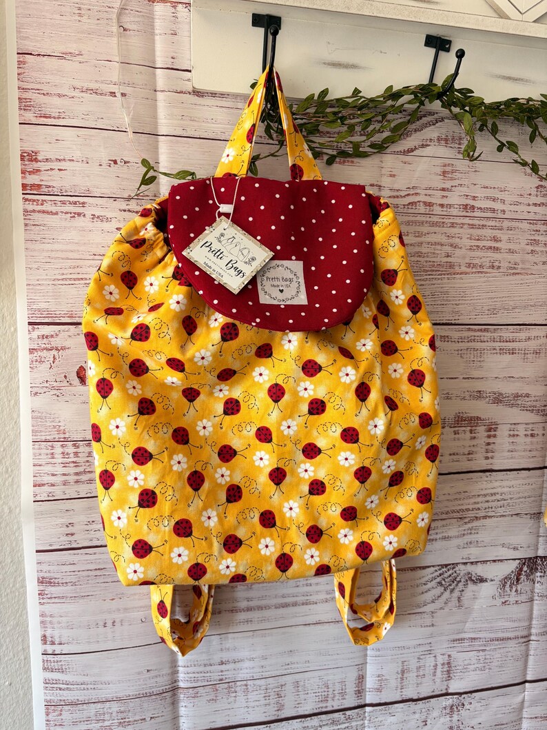 Peut inclure: Un sac &agrave; dos &agrave; cordon en tissu jaune avec un motif de coccinelles rouges et de marguerites blanches. Le sac poss&egrave;de un rabat rouge fonc&eacute; &agrave; pois blancs et une &eacute;tiquette indiquant Pretti Bags Made in USA. Il comprend deux anses et des bretelles.