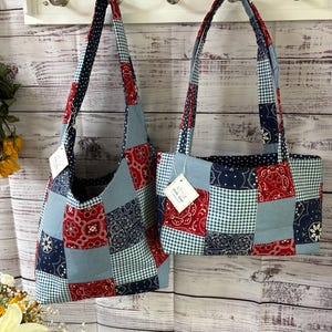 Puede incluir: Dos bolsos tote de patchwork con correas a juego cuelgan de ganchos. Las bolsas presentan una mezcla de telas rojas, blancas y azules en varios patrones, incluyendo cachemir, cuadros y lunares. Uno es un bolso de hombro, el otro un tote rectangular.
