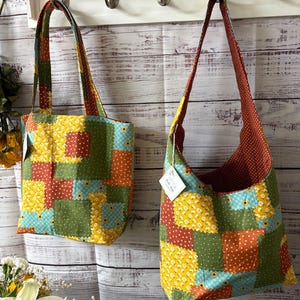 Könnte beinhalten: Zwei Patchwork-Tragetaschen mit langen Trägern hängen an einer weißen Holzwand. Die Taschen haben ein farbenfrohes Patchwork-Design mit Quadraten aus gelben, grünen, orangefarbenen und blauen Stoffen. Eine ist eine Tragetasche, die andere eine Umhängetasche.