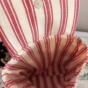 Peut inclure: Un sac en tissu avec une ouverture sur le dessus et un rabat, pr&eacute;sentant un motif &agrave; rayures verticales rouges et blanches. L'int&eacute;rieur est doubl&eacute; du m&ecirc;me tissu ray&eacute; rouge et blanc. Un bouton-pression magn&eacute;tique argent&eacute; est fix&eacute; au rabat.