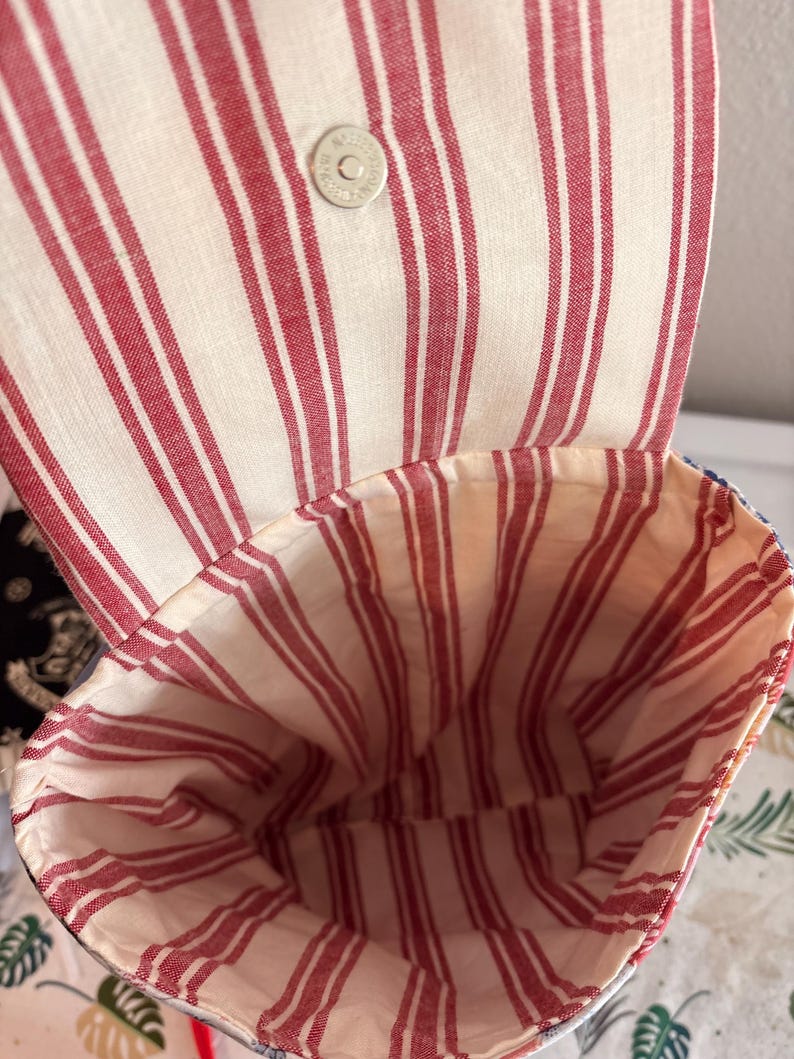 Peut inclure: Un sac en tissu avec une ouverture sur le dessus et un rabat, pr&eacute;sentant un motif &agrave; rayures verticales rouges et blanches. L'int&eacute;rieur est doubl&eacute; du m&ecirc;me tissu ray&eacute; rouge et blanc. Un bouton-pression magn&eacute;tique argent&eacute; est fix&eacute; au rabat.