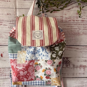 Peut inclure: Un sac &agrave; dos en patchwork compos&eacute; de divers motifs floraux, vichy et ray&eacute;s dans des tons de rouge, bleu, vert et jaune. Le rabat sup&eacute;rieur pr&eacute;sente des rayures verticales rouges et blanches avec une &eacute;tiquette blanche indiquant Pretti Bags Made in USA.