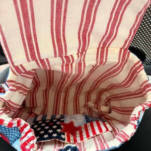 Peut inclure: Un sac &agrave; dos en tissu avec une ouverture sur le dessus, dot&eacute; d'une doublure int&eacute;rieure &agrave; rayures verticales cr&egrave;me et rouge. L'ext&eacute;rieur pr&eacute;sente un motif d'&eacute;toiles bleues et blanches avec une &eacute;toile en tissu rouge sur la poche avant.
