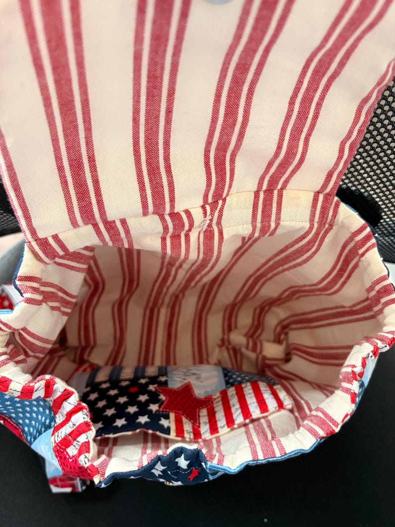 Peut inclure: Un sac &agrave; dos en tissu avec une ouverture sur le dessus, dot&eacute; d'une doublure int&eacute;rieure &agrave; rayures verticales cr&egrave;me et rouge. L'ext&eacute;rieur pr&eacute;sente un motif d'&eacute;toiles bleues et blanches avec une &eacute;toile en tissu rouge sur la poche avant.