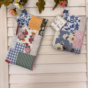 May include: Two rectangular fabric pouches featuring patchwork designs with floral, gingham, and striped patterns in blue, white, pink, green, and yellow. Each pouch has a zipper closure, a metal ring, and a tag labeled Pritti Bags Made in USA.