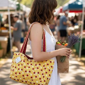 May include: A yellow tote bag featuring a repeating pattern of red cherries and small white flowers. The bag has two shoulder straps in a deep red fabric with a white polka dot print. A small white rectangular tag is attached to the side.