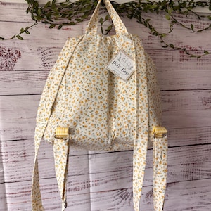 Peut inclure: Un sac &agrave; dos en tissu avec une base blanche et un petit motif floral jaune et vert. Le sac poss&egrave;de deux bretelles, une poign&eacute;e sup&eacute;rieure et une &eacute;tiquette blanche attach&eacute;e portant l'inscription Pretti Bags Made in USA avec un dessin.