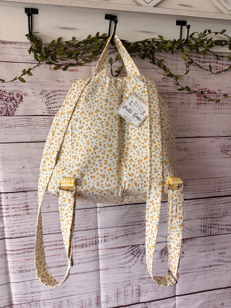 Peut inclure: Un sac &agrave; dos en tissu avec une base blanche et un petit motif floral jaune et vert. Le sac poss&egrave;de deux bretelles, une poign&eacute;e sup&eacute;rieure et une &eacute;tiquette blanche attach&eacute;e portant l'inscription Pretti Bags Made in USA avec un dessin.