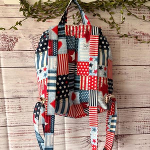 Peut inclure: Un sac &agrave; dos en tissu avec un motif patchwork aux couleurs rouge, blanc et bleu. Le design comprend des &eacute;toiles, des rayures et des c&oelig;urs. Il poss&egrave;de deux bretelles r&eacute;glables et une poign&eacute;e sup&eacute;rieure, fabriqu&eacute;s dans le m&ecirc;me tissu &agrave; motifs.