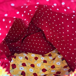 Peut inclure: Un sac &agrave; cordon en tissu avec un ext&eacute;rieur rouge vif &agrave; pois blancs. La doublure int&eacute;rieure est en tissu jaune contrastant, orn&eacute; d'un motif r&eacute;p&eacute;titif de petites coccinelles rouges et de fleurs de marguerites blanches sur toute la surface.