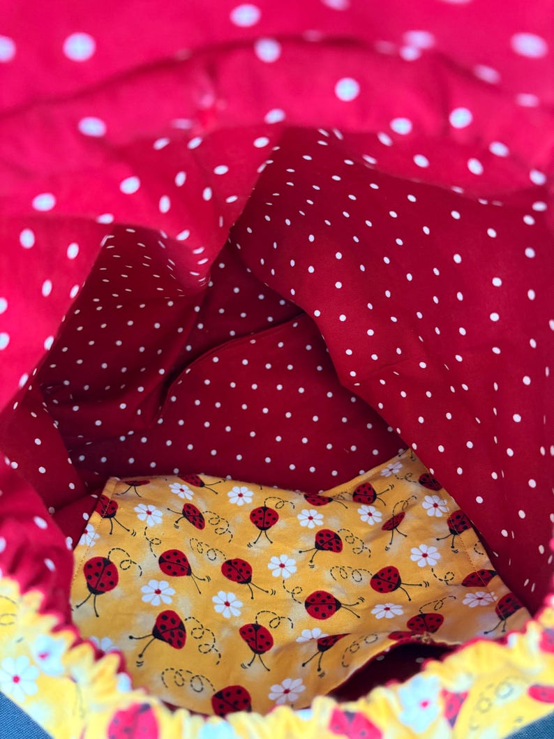 Peut inclure: Un sac &agrave; cordon en tissu avec un ext&eacute;rieur rouge vif &agrave; pois blancs. La doublure int&eacute;rieure est en tissu jaune contrastant, orn&eacute; d'un motif r&eacute;p&eacute;titif de petites coccinelles rouges et de fleurs de marguerites blanches sur toute la surface.