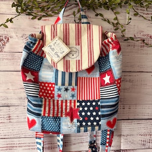 Peut inclure: Un sac &agrave; dos en tissu avec un motif patchwork compos&eacute; de rayures, d'&eacute;toiles et de c&oelig;urs rouges, blancs et bleus. Le rabat sup&eacute;rieur porte une &eacute;tiquette indiquant Pretti Bags Made in USA ainsi qu'une &eacute;tiquette assortie sur le devant.