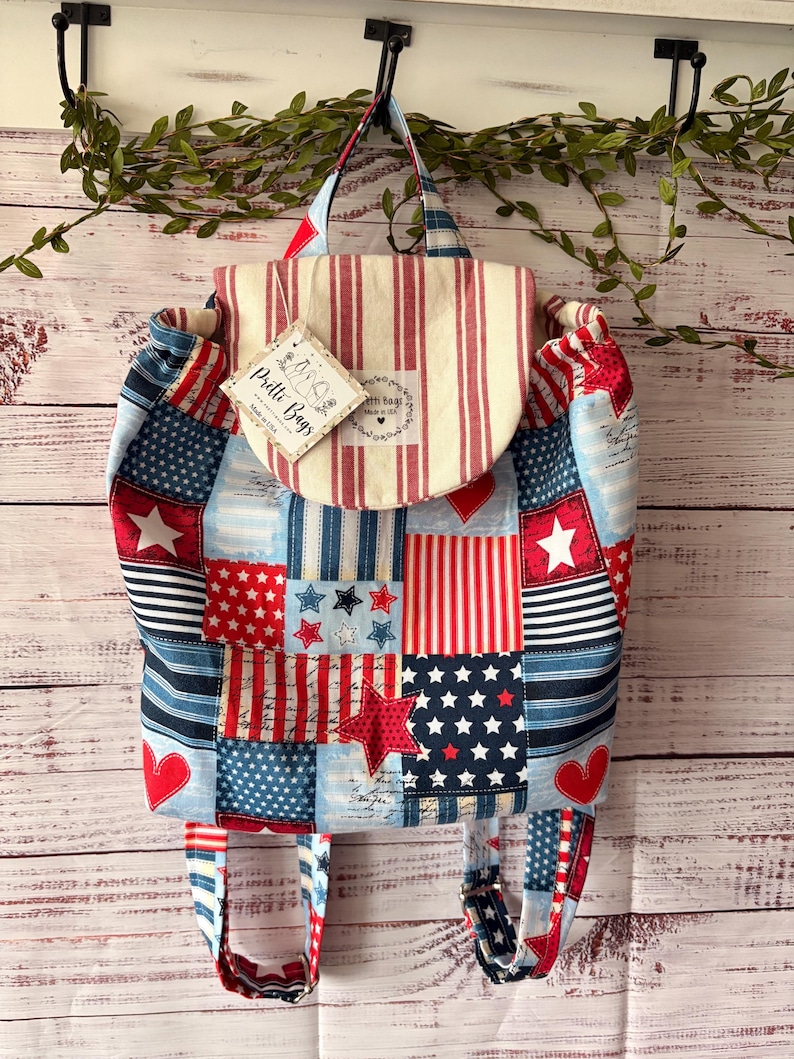 Peut inclure: Un sac &agrave; dos en tissu avec un motif patchwork compos&eacute; de rayures, d'&eacute;toiles et de c&oelig;urs rouges, blancs et bleus. Le rabat sup&eacute;rieur porte une &eacute;tiquette indiquant Pretti Bags Made in USA ainsi qu'une &eacute;tiquette assortie sur le devant.
