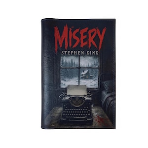 Könnte beinhalten: Ein rechteckiger Buchumschlag mit dem Titel MISERY in gezackten roten Buchstaben über dem Autorennamen Stephen King. Die Grafik zeigt eine schwarze Schreibmaschine auf einem Holztisch vor einem Fenster mit einer verschneiten Landschaft.