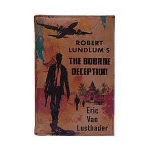 Puede incluir: Una funda de libro de cuero marrón con el título "The Bourne Deception" de Robert Lundlum y Eric Van Lustbader. La portada incluye la silueta de un hombre con una pistola, un avión y una escena tropical.