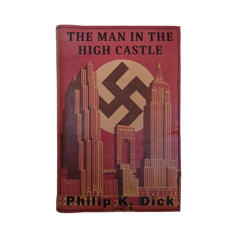 K&ouml;nnte beinhalten: Ein rotes, ledergebundenes Buch mit dem Titel "The Man in the High Castle" und dem Namen des Autors "Philip K. Dick". Der Umschlag zeigt ein stilisiertes Bild einer Skyline mit einem gro&szlig;en Hakenkreuz in der Mitte.