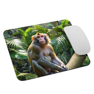 Puede incluir: Alfombrilla de ratón rectangular con un mono sentado en una rama de árbol, rodeado de vegetación verde. Un ratón de ordenador blanco está a la derecha. El mono tiene pelaje marrón claro y una expresión concentrada. Accesorio de ordenador.