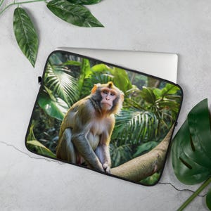 Puede incluir: Funda para portátil con un mono sentado en una rama de árbol, sobre un fondo de exuberante follaje verde. El mono tiene pelaje marrón claro y una expresión concentrada. La funda es negra y protege un portátil.